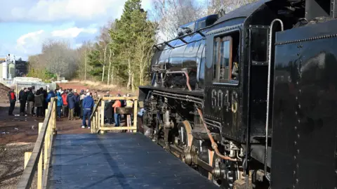 Churnet Valley Railway Viewed from the side, a black locomotive is stopped against a makeshift wooden platform. A group of people are stood slightly away from the platform, in front of the engine. There are trees in the distance.