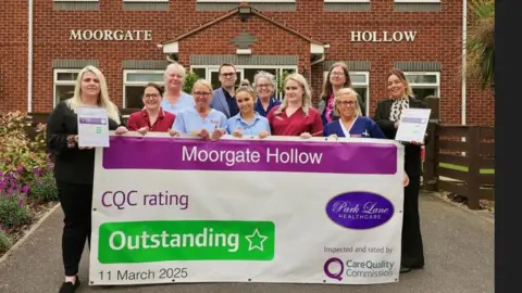 An image of 11 people standing in front of a red brick building. There are 10 women and one man. Some of the women are dressed in nursing tunics. They are holding a giant banner which reads Moorgate Hollow, CQC rating Outstanding, 11 March 2025.