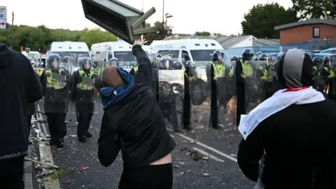 Getty Images A group of people, pictured from behind, face off against riot police holding shields. A person in the centre of the image is throwing a what appears to be a wooden planter in the direction of the officers. 