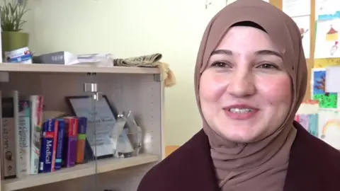 A woman in a pale brown head scarf and maroon top looks at the camera. She is sitting in an office. To her right is a glass-fronted cabinet, with books inside and a plant on top of it. The wall is pale yellow in colour.