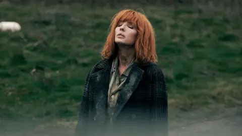 Sky Kelly Reilly standing in a field, looking to the sky with her eyes closed. She has copper shoulder length hair with a fringe. She wears a green shirt and a green tartan coat.