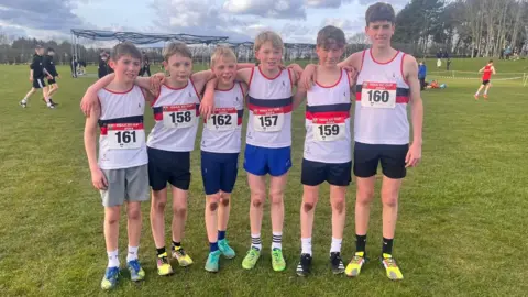 Taunton School A group of boys stand in height order with their arms wrapped around each other's shoulders. They are all wearing white tank tops with race numbers attached to their tops. They are wearing shorts, socks and running shoes.