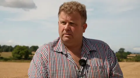 William Maughan, a middle aged man with blonde, curly hair. He is stood in the middle of a harvested wheat field, which stretches out into a backdrop of rolling, beige hills under a light blue sky.