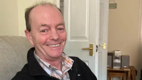 BBC Mark Abel at home, sitting on a sofa, wearing a shirt and black jumper. He is smiling at the camera and looking happy and excited.