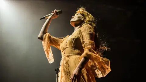Getty Images Florence Welch wears a cream flowy dress as she performs on stage. She is singing in to a microphone as a spotlight beams down on her. 