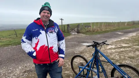 Matt Hodgson, wearing his GB jacket, standing with the dry land sled, which has four wheels and an aluminium frame.