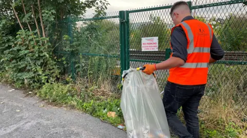 Man in a bright orange vest carrying a bag full of rubbish along a street