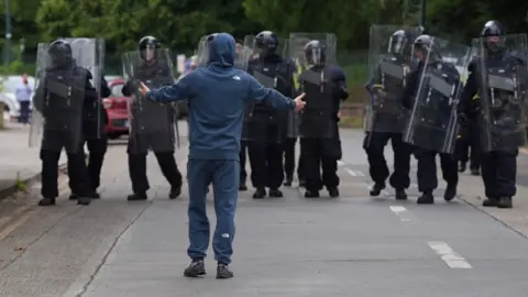 PA Mediaman in hoodie, armen uitgestrekt voor de lijn van de Ierse oproerpolitie