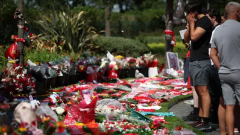 A crowd of people, including a man with his face in his hands, stand on the edge of a grassy area blanketed in flowers, football shirts, scarves and balloons