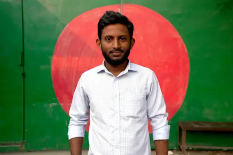 A young man wearing a white shirt stands in front of a wall on which is painted a big red circle, with a bright green background.