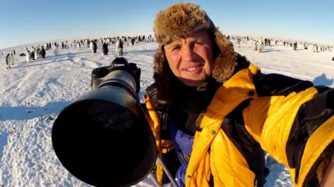 a man in a yellow jacket and furry hat with a large camera standing on a stretch of snow with hundreds of penguins in the background
