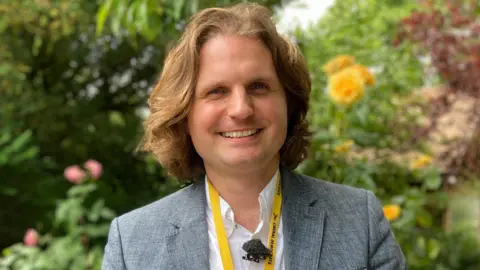 Alex Dunlop/BBC Steffan Aquarone is standing outside in front of green trees and bushes. He is looking directly at the camera and smiling. He is wearing a grey suit jacket and a white shirt.
