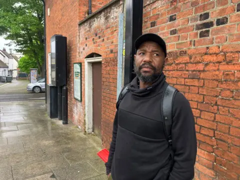 Matt Marvel/BBC Ignatius is standing on a pavement dressed all in black. He is standing in front of a brick wall.