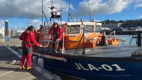 BBC Lifeboat crews in red are tying up a bright orange and blue lifeboat to port. The boat is labelled as 'JLA-01'. 