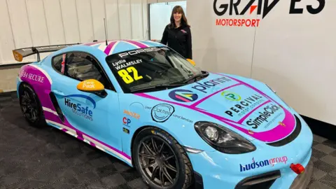 Lydia Walmsley dressed in all black stands in a car show room with her blue Porsche.