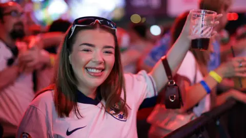 Getty Images A girl wholes a beer. She is wearing a England football show. 