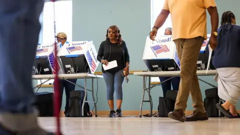 People casts ballots during early voting at the Garners Ferry Adult Activity Center on 26 May 2026 in Hopkins, South Carolina. A woman in a black top and gray leggings walks between two voting tables, holding a white piece of paper and her phone
