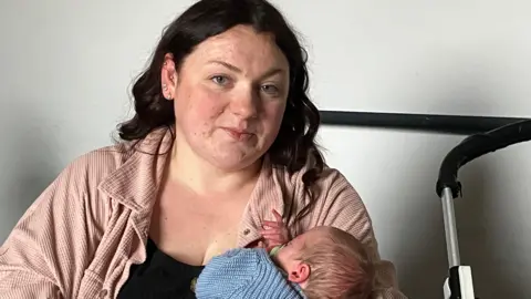 A woman, aged 30, looks to camera. She has dark hair and blue eyes and is wearing a black vest and pale pink shirt / jacket over the top. She is cradling her 6 week old baby son, who is wearing a pale blue knitted cardigan