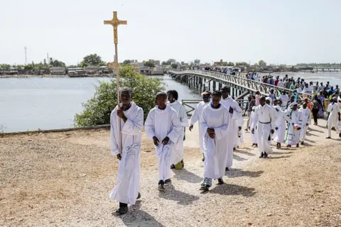 Patrick Meinhardt / AFP / Getty Images An altar boy in white carries a cross as he leads the procession to the cemetery  on the island of Fadiouth - Saturday 1 November 2025.