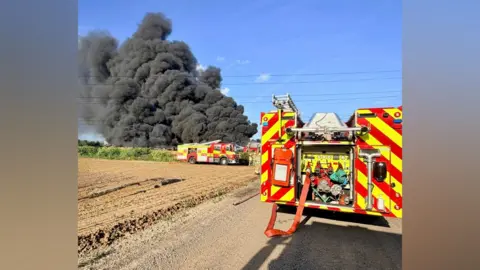 Essex County Fire and Rescue Service A cloud of thick black smoke in an otherwise blue sky. In the foreground are a ploughed field and fire engines.