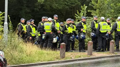 Nadira Tudor/BBC A large group of police officers, more than 15, are stood on a public path near a rain soaked road, many are holding police helmets in their hands. All are wearing high-vis vests.