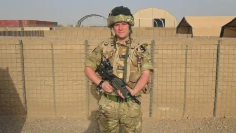 Combat Stress A photo of a young man in Afghanistan wearing a brown and green camo outfit with a matching helmet. There is a black gun in his hands. Behind him is a sandy wall with metal grating over it, with brown air base type buildings in the background