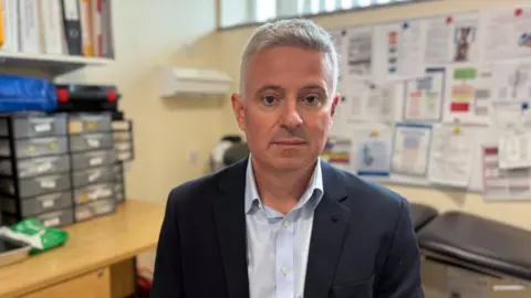 STEVE HUBBARD/BBC Paul Scott wearing a black suit and a pale blue shirt. He has grey hair and is sitting in a trust office. There are lots of metrics pinned to the wall behind him.