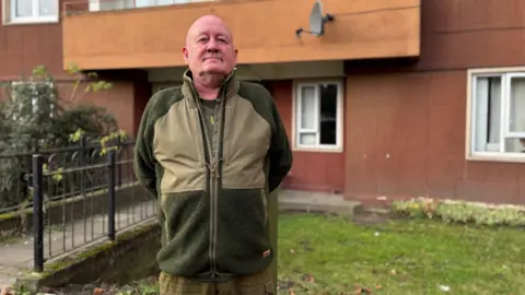 John Butcher wearing a two-tone green fleece over a green t-shirt and green trousers. He is bald. He is standing in front of Westfield Court.