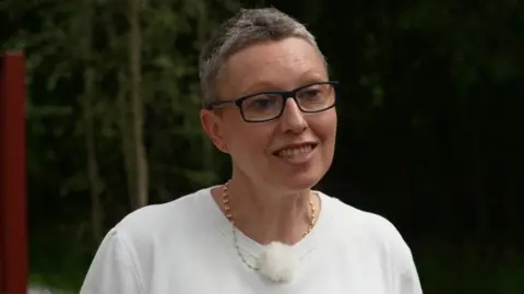Georgina Baines has short hair and is wearing a white top, dark framed glasses and a gold necklace. She is standing in a park area speaking to a TV reporter.