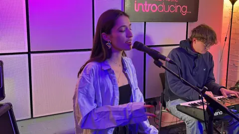 Nina Arya sits on a stool in a studio and is singing into a microphone. She wears an over-sized blue shirt. A man with glasses plays a keyboard. There is a purple light glowing and the BBC Introducing sign is on a wall.