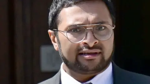 Wales News Service Ragu is standing outside Cardiff crown court. He has short dark hair and a cropped beard. He is wearing a suit and round metal rimmed glasses