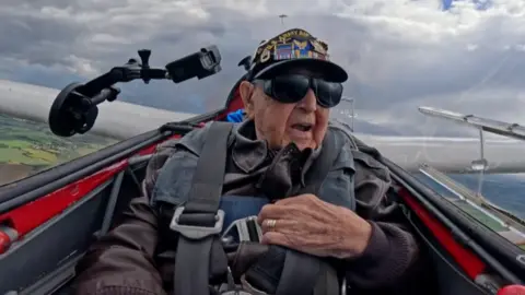 Casey Bukowski is wearing a leather jacket strapped into a plane seat, with clouds behind him and some fields visible below.