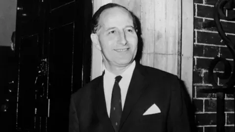 Getty Images Black and white photograph of Terence O'Neill, he has short hair and is smiling to the side. Wearing a suit jacket, shirt and tie. Pictured outside a bricked building.