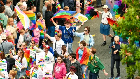Pacemaker A crowd of people taking par and watching a parade. People are dressed in bright colours and many of them are holding posters and flags. 