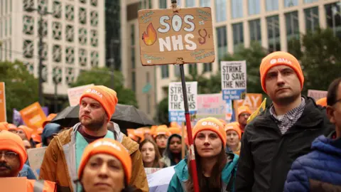 ADAM VAUGHAN/EPA-EFE/REX/Shutterstock People in orange hats with BMA written on them gather in Manchester. A woman in the middle is holding a sign that says 'SOS NHS'