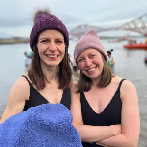 Two women in black swimming costumes and purple beanie hats stand in the Firth of Forth, smiling at the camera.