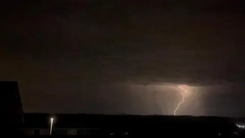 Izzy A bolt of lightning comes down from the night sky in the distance, viewed from a residential estate.