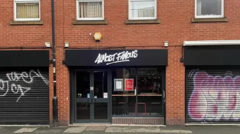 A black shopfront for Almost Famous, set underneath some windows on a redbrick building, in between two shuttered shopfronts. 