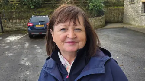 A lady in a navy blue rain coat stands in front of a parked car. The walls of a house are seen in the background. She has shoulder length brown hair.