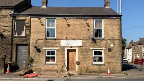 Image shows a dilapidated pub on the end of a street. It is built of stone and its front door is boarded over. 