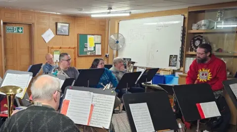 Ilkeston Brass Five men and one woman are sat in an old golf pavilion in front of black music stands. Some of them can be seen holding brass instruments and there is sheet music on the stands.