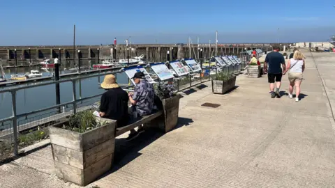 Two people in sun hats sitting on a bench overlooking a harbour in warm weather. Others are walking past wearing shorts and t-shirts. Boats can be seen on the harbour with information boards along the railings.