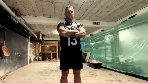 Garry Bell, standing with crossed arms and wearing shorts and a T-shirt, inside Contender Gym which is dirty and looks like it's in the process of being rebuilt, with no proper floor covering, plastic sheeting covering bits of gym kit, raw wood frames and what looks like a bath for mixing concrete in the background.