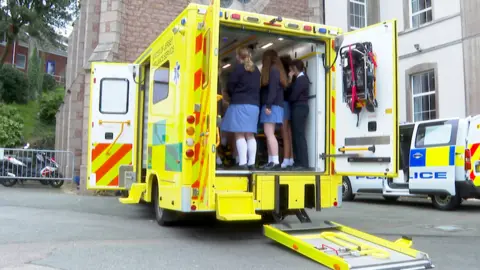 BBC A yellow ambulance with students in it looking around, parked on a Tarmac car park with buildings in front, a police van on the right beside it
