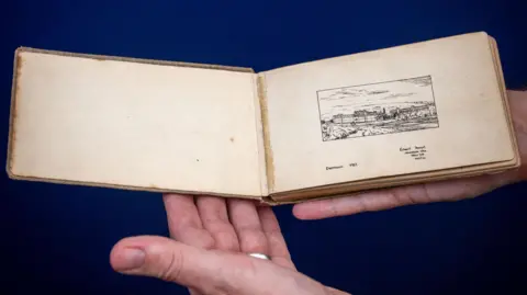 HANSONS AUCTIONEERS A hand holding the album. It is brown in colour with a image pictured on the right page. The person holding the book is wearing a silver ring. The album looks delicate and is only slightly larger than the person's hand.