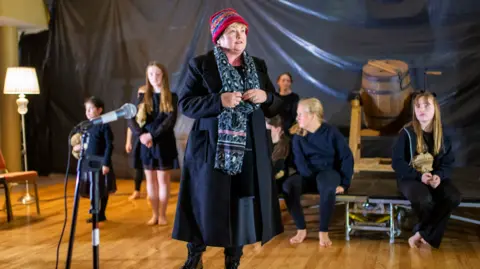 Cuimhneamh Social Media A school play is taking place up at the front is a woman with a hat, black coat and blue patterned scarf. In the background is children wearing a navy top and trousers, or a dress while sitting on a stage.