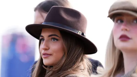 A young woman with long brown hair and a brown trilby-style hat at the Cheltenham Festival 2026. 