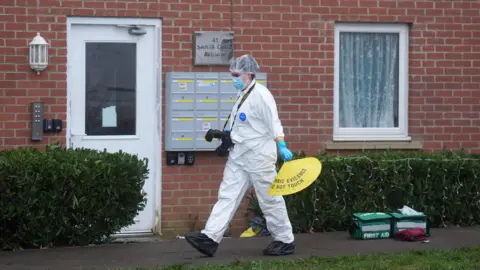 PA Media A forensics officer, in a white suit, hair net and face mask, is walking from right to left outside terraced housing. They have a camera and are holding a yellow cone. There are two green first aid bags on the pavement outside the houses.