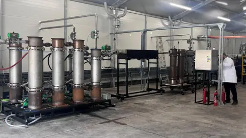 A large mechanical device with tall stacks connected to other equipment installed in a warehouse being operated by a man in a white coat