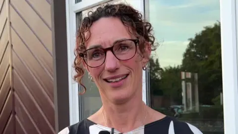 Sarah Sanderson/BBC A woman wearing a black, white and beige patterned top has a small microphone attached to it and is looking slightly to the right smiling. She has curly brown hair and is wearing glasses and is stood in front of a house.